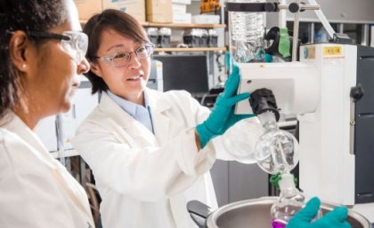 Two female scientists in whitecoats, gloves and goggles inspect a same from a machine.